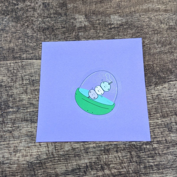 3 Colorful Dango Mochi on a stick inside a gachapon style toy capsule in the form of a sticker with see through vinyl  on a purple post it note to show qualities of clear vinyl portions of sticker